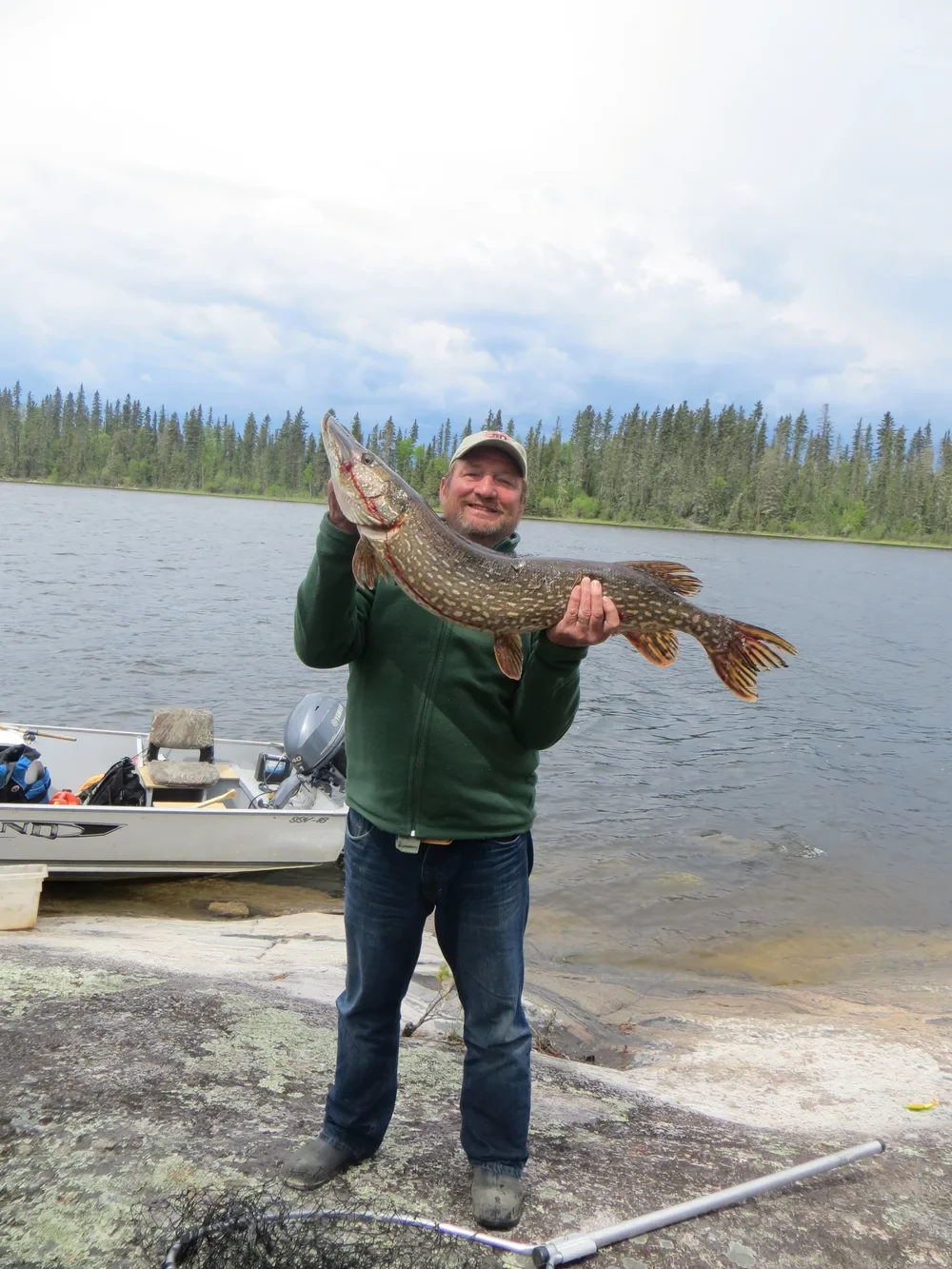Man holding a large pike on rocky shoreline, boat behind him