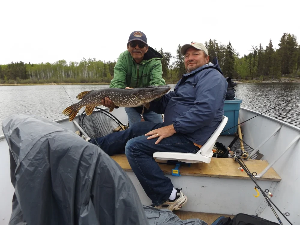 Two men in a boat, one holding a large Northern Pike, overcast sky