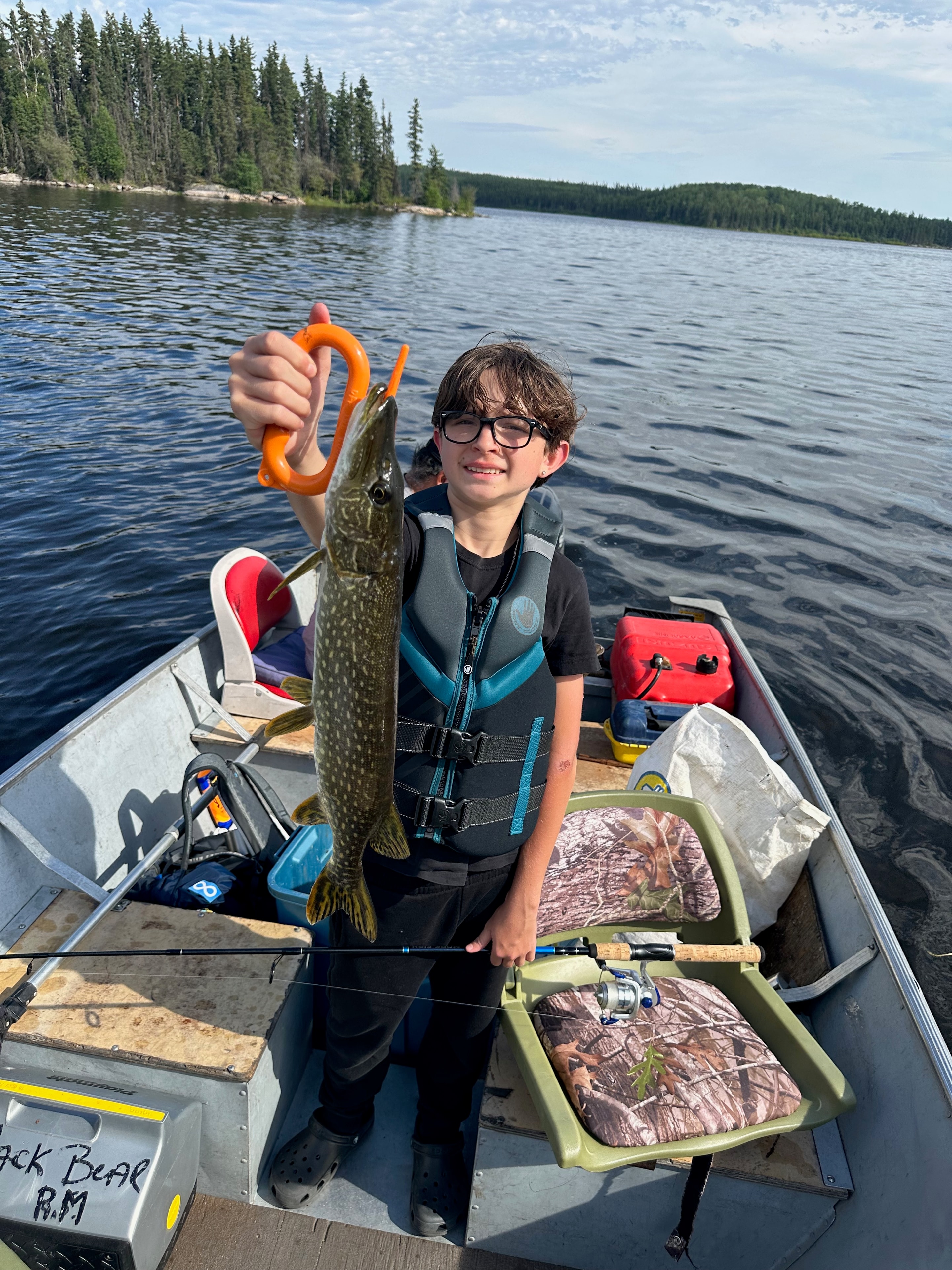 Boy in MFFL hat holding a walleye with a lip gripper on a boat, lake and boreal forest backdrop