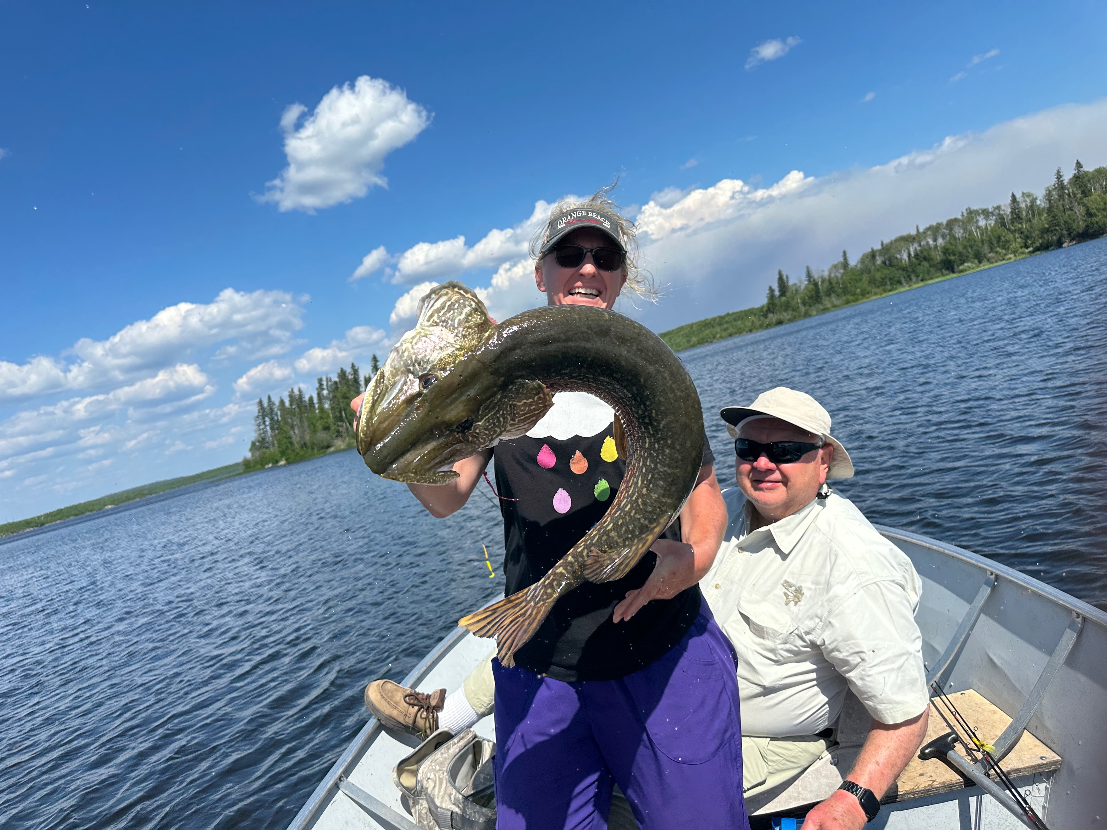 Woman holding a massive curled Northern Pike in a boat with a man seated behind her, sunny lake
