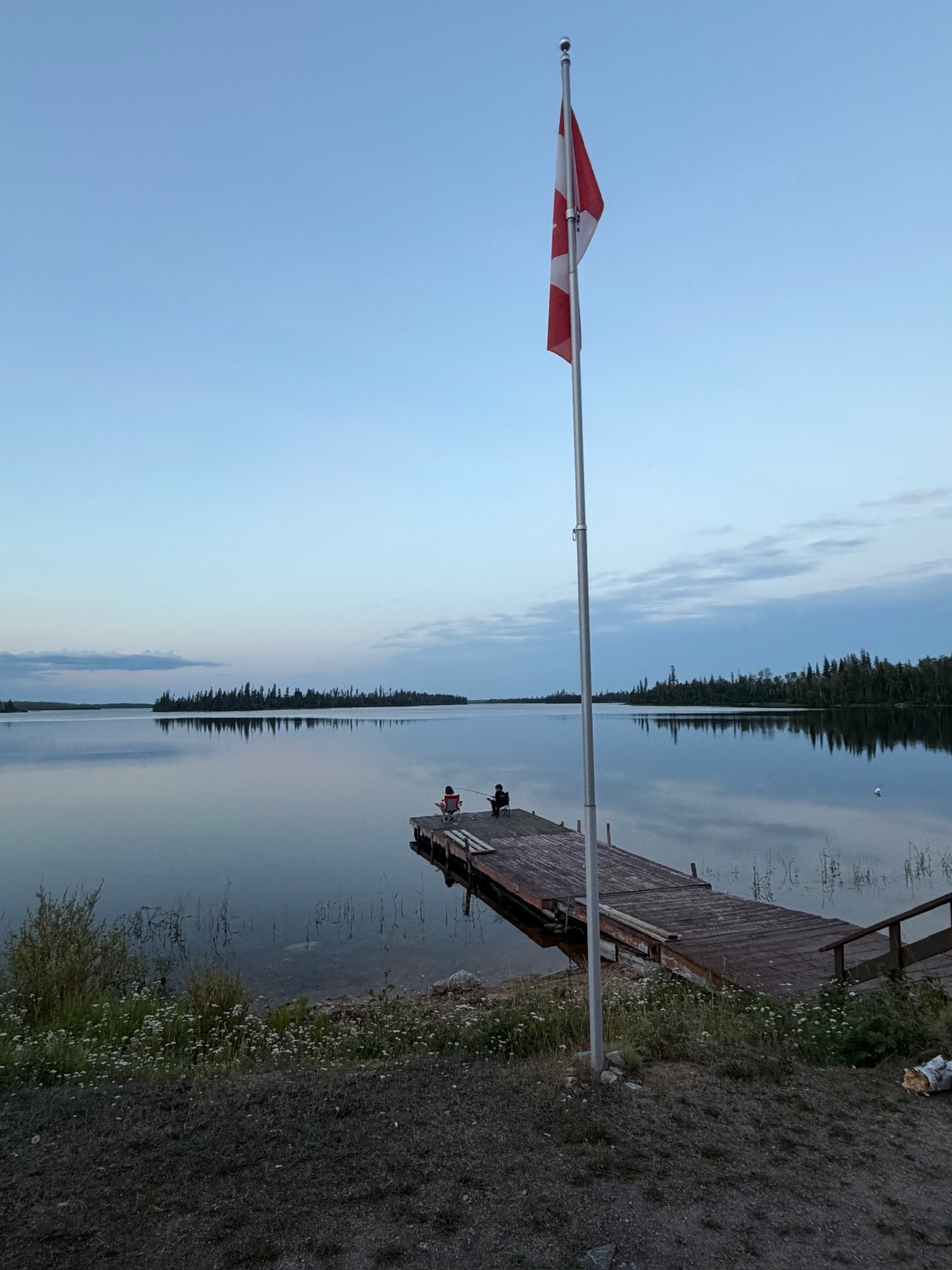 Canadian flag on dock with two people fishing at dusk, calm lake reflection