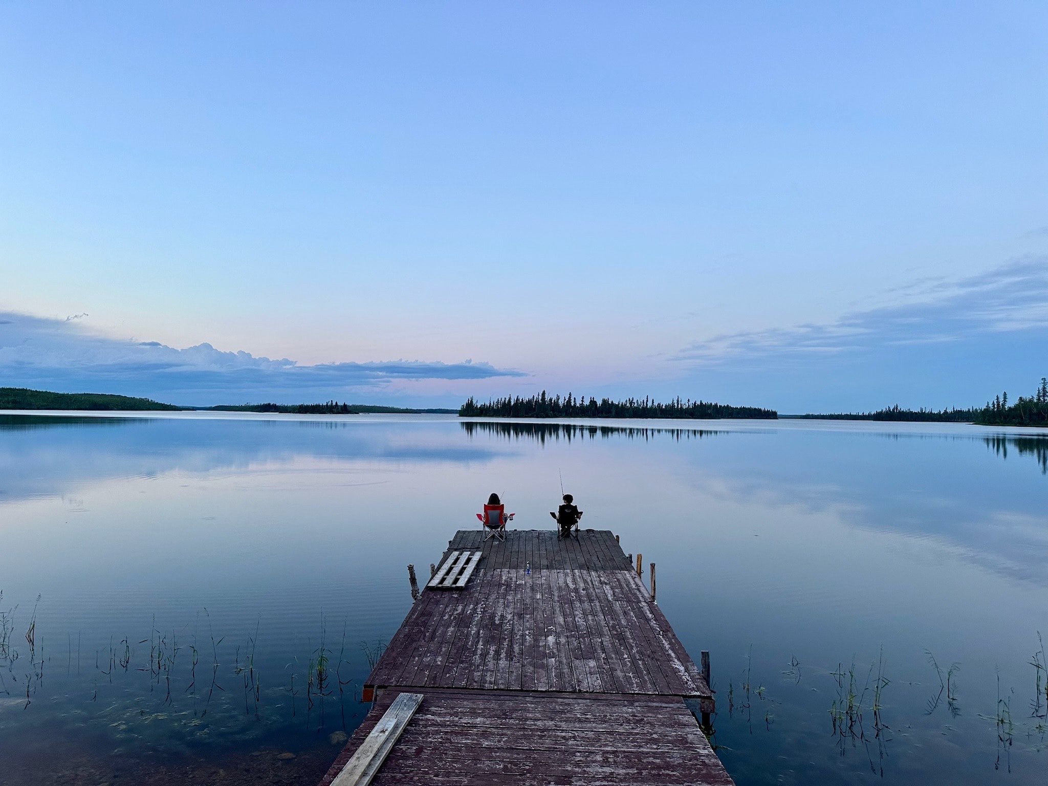 Two people sitting in camp chairs fishing from the dock at dusk, lake perfectly still